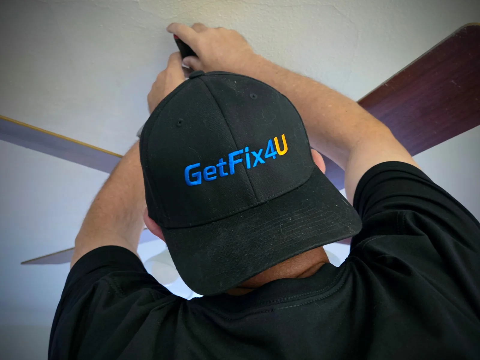 Technician adjusting a ceiling fan wall control while wearing a GetFix4U cap.