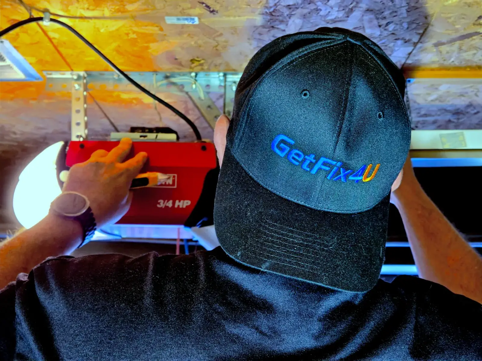 Technician adjusting a garage door opener unit in a garage with mixed work-light and daylight.